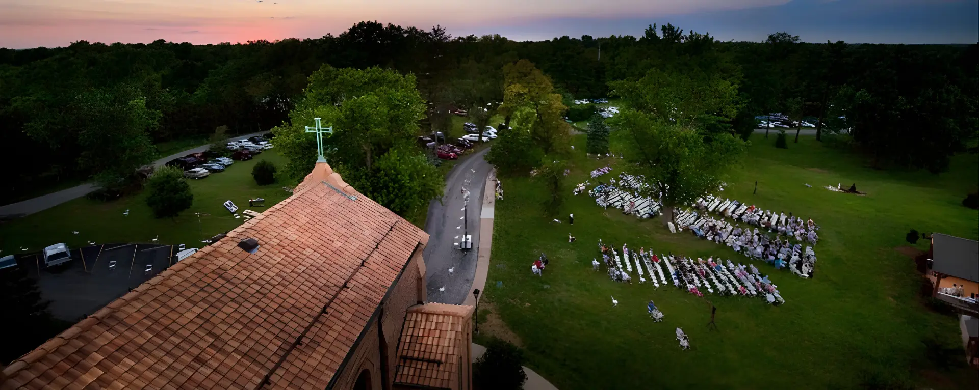 Aerial view of a church with a cross on the roof, surrounded by trees at sunset. Nearby, people gather outside on chairs arranged in rows, suggesting an outdoor event. The scene conveys a peaceful, communal atmosphere.