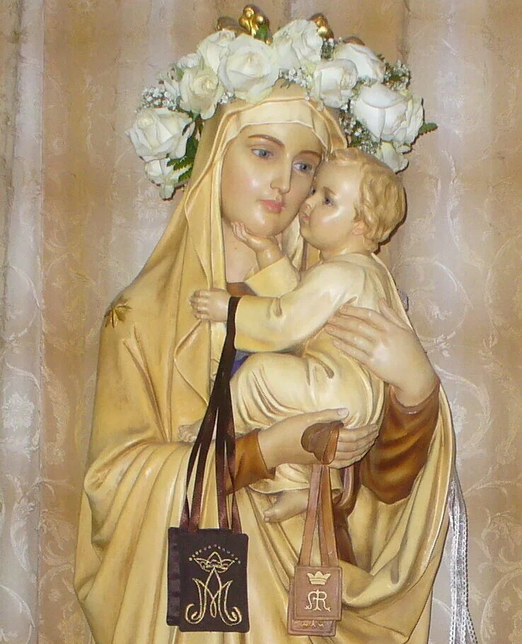 A statue of Mary holding baby Jesus, adorned with a crown of white roses. She wears a golden robe and holds two brown scapulars, exuding serenity and devotion.