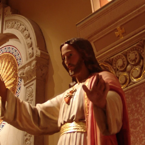 Statue of Jesus with arms outstretched, adorned in gold and red robes, set in a decorative church interior. White floral design overlay on the left.