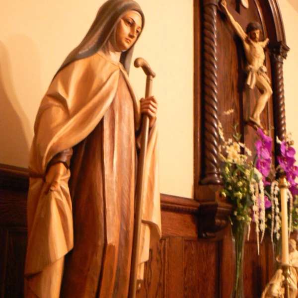 Statue of Jesus with arms outstretched, adorned in gold and red robes, set in a decorative church interior. White floral design overlay on the left.