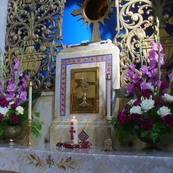 An ornate altar with golden details, surrounded by pink and white flowers. A candle with a red cross is placed in front, conveying a solemn tone.