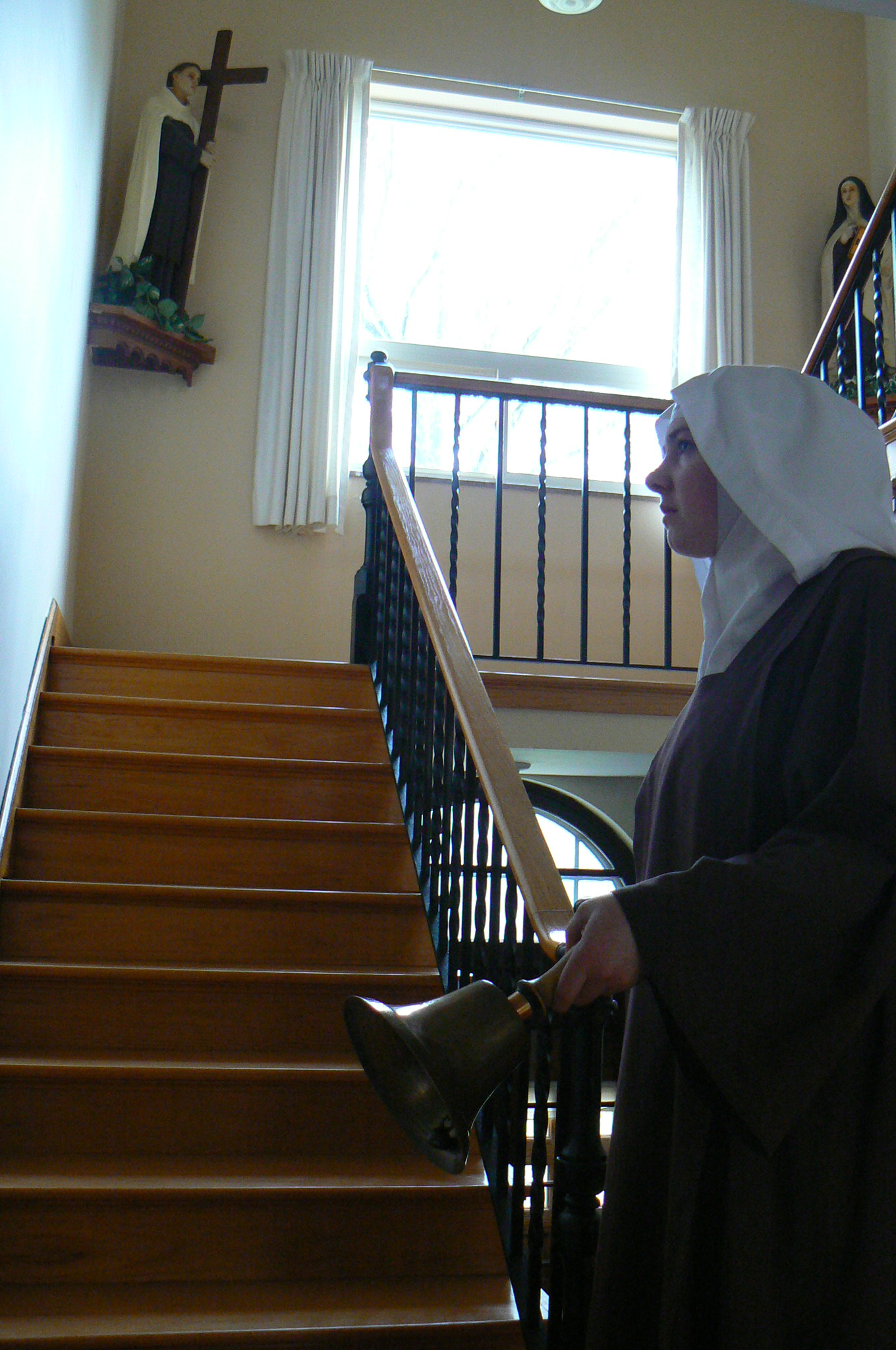 A nun in a brown habit and white veil stands by a wooden staircase, holding a bell. She gazes up at a saint statue. The scene is peaceful and solemn, with floral designs on the left.