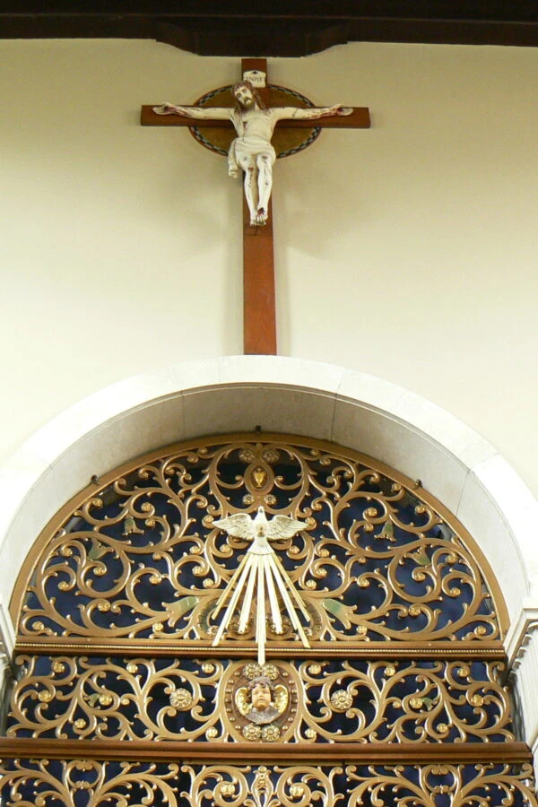 Crucifix on a wall above an ornate gold and blue decorative grill, featuring intricate scrollwork and a dove motif, conveying a solemn and reverent tone.