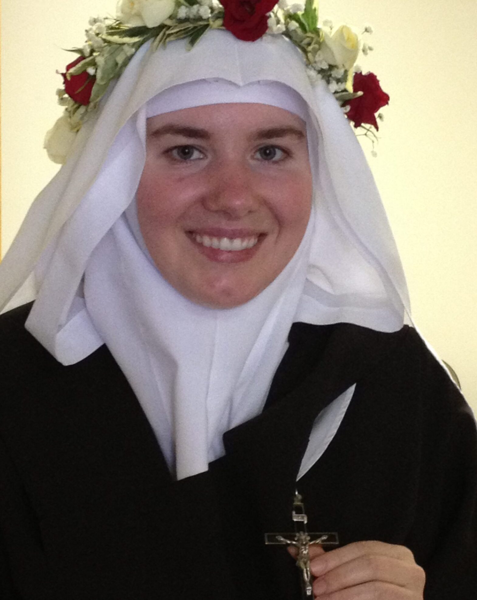 A smiling woman wearing a white habit and a floral crown with red and white flowers. The background is simple, and there's a floral illustration overlay on the left.