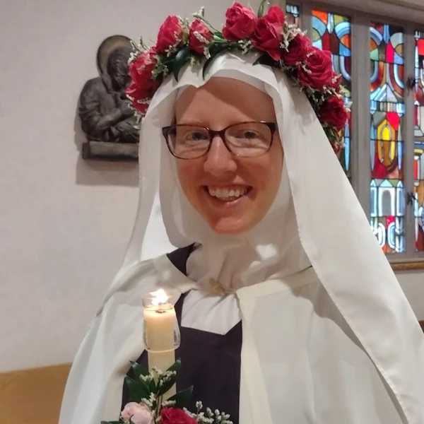 A smiling person in a white religious habit and glasses, adorned with a floral crown and holding a lit candle. Stained glass and floral sketch frame the scene.