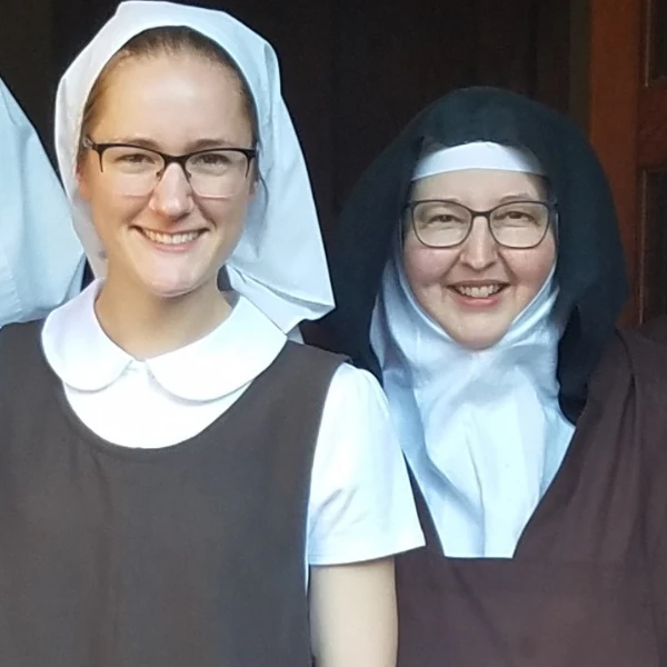 Two women wearing religious habits smile warmly. One wears a white headdress, the other a black one. Floral designs accent the left side.