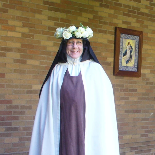 A joyful nun wearing a white floral crown, brown habit, and white mantle stands against a brick wall with a religious painting, exuding serenity. Floral design frames the left.