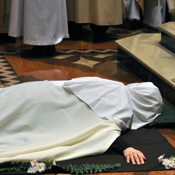 A person in religious attire lies prostrate on a church floor, suggesting a solemn ceremony. A floral border adds to the reverent atmosphere.
